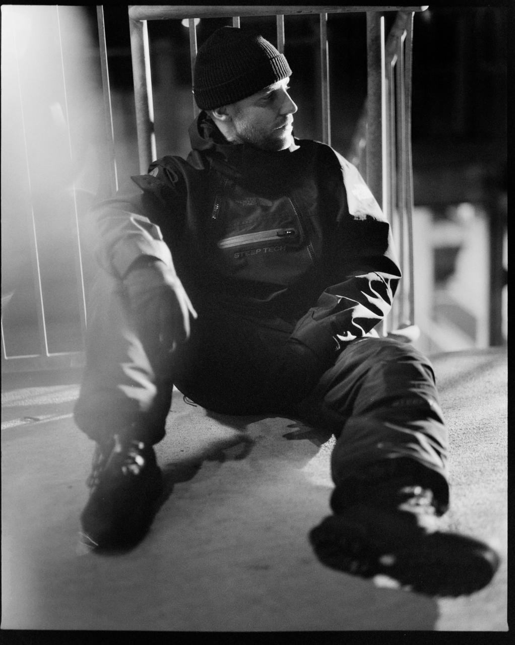 Man seated on ground, wearing a beanie and jacket with text, against a metal structure