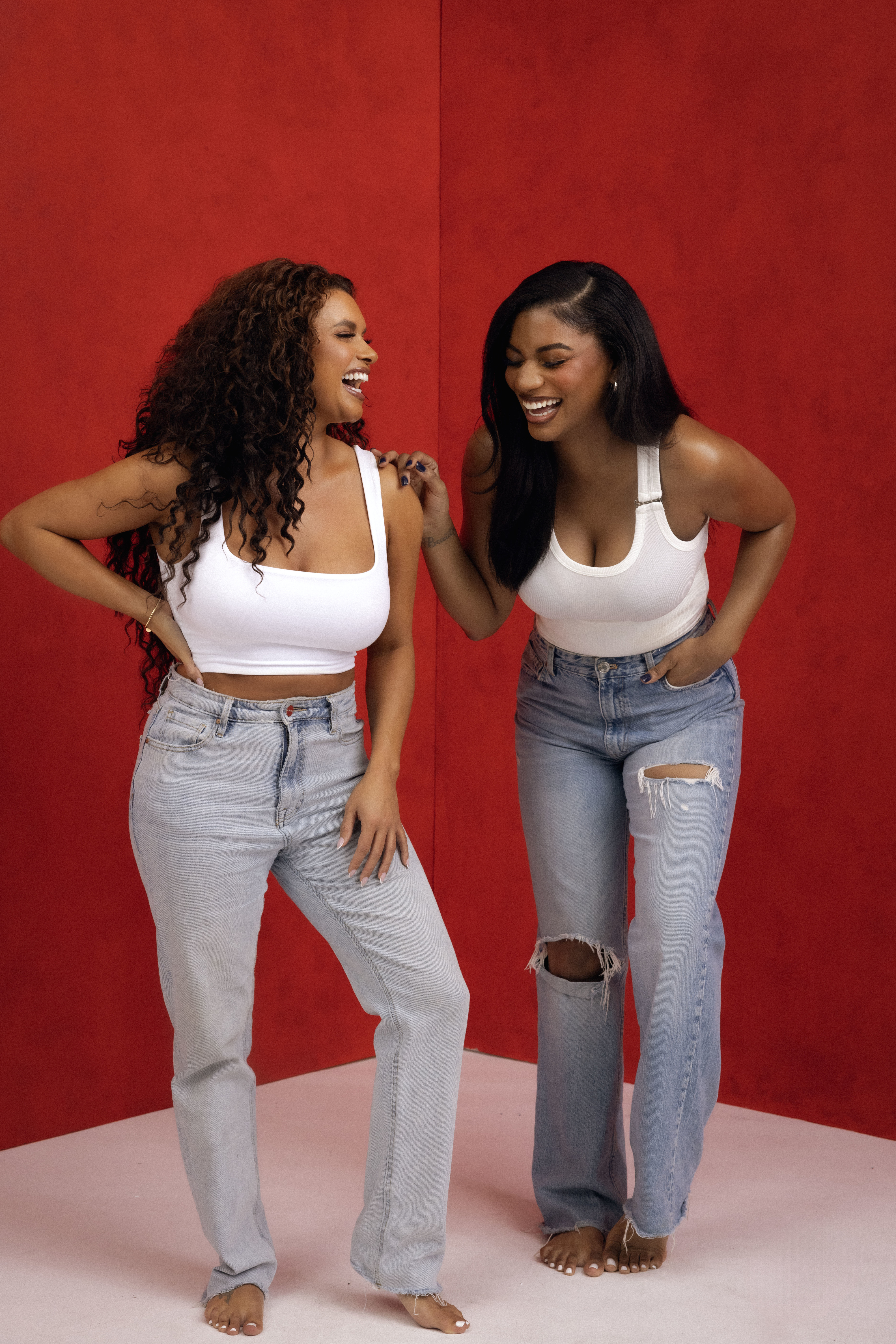 Two women in white tops and jeans smiling and posing playfully against a red backdrop