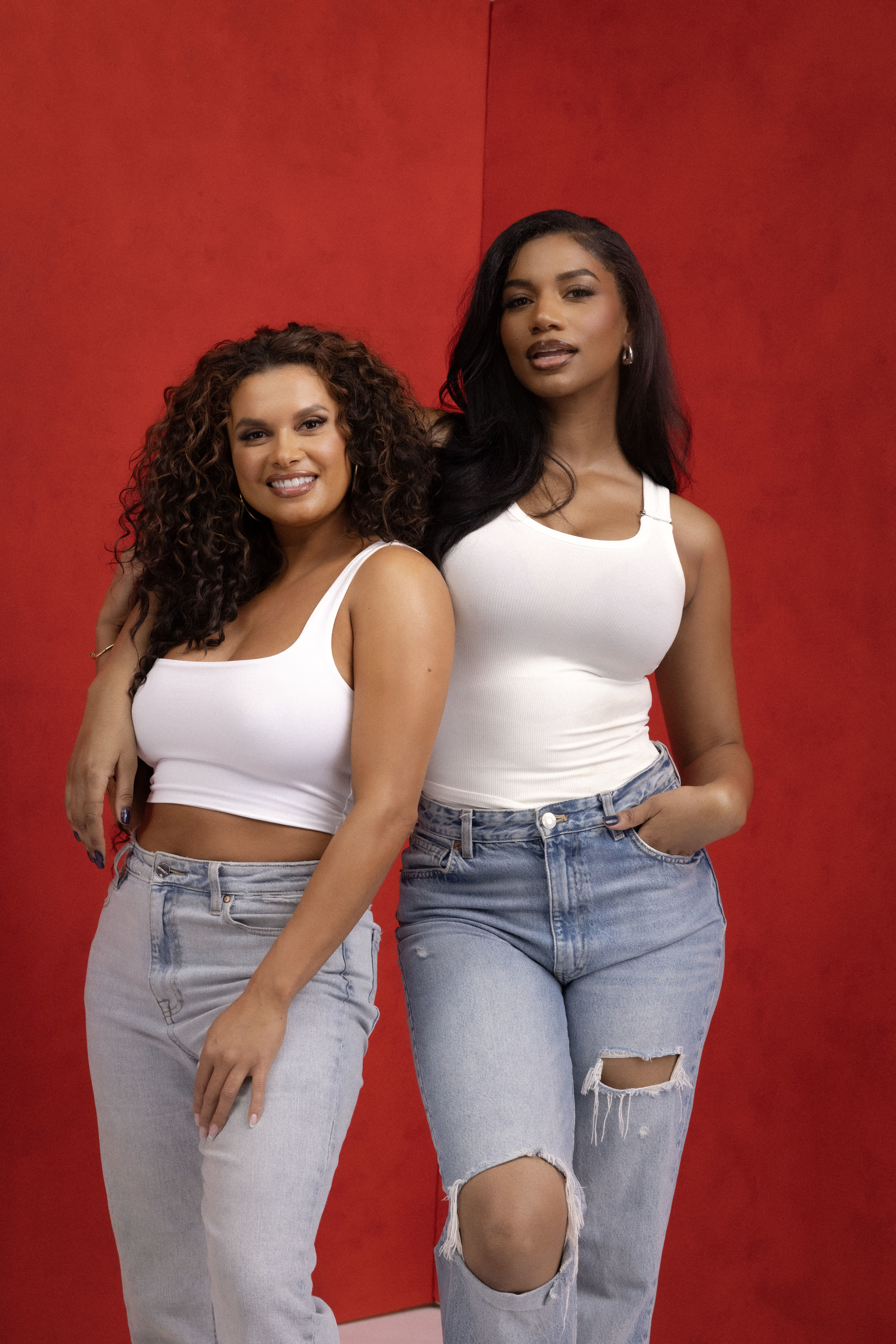 Two women posing in casual white tops and jeans against a red backdrop