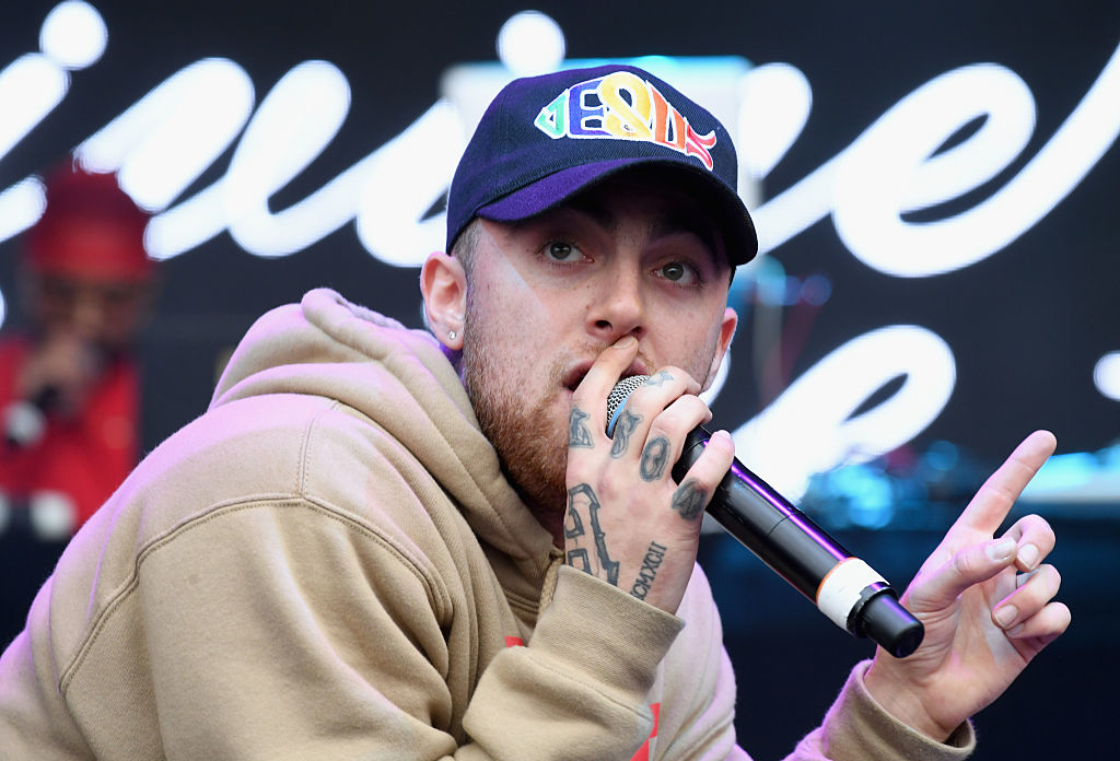 Mac Miller performing on stage, wearing a hoodie and baseball cap, holding a microphone