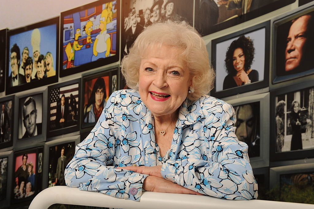 Betty White smiles in front of a wall with framed photos, wearing a floral blouse