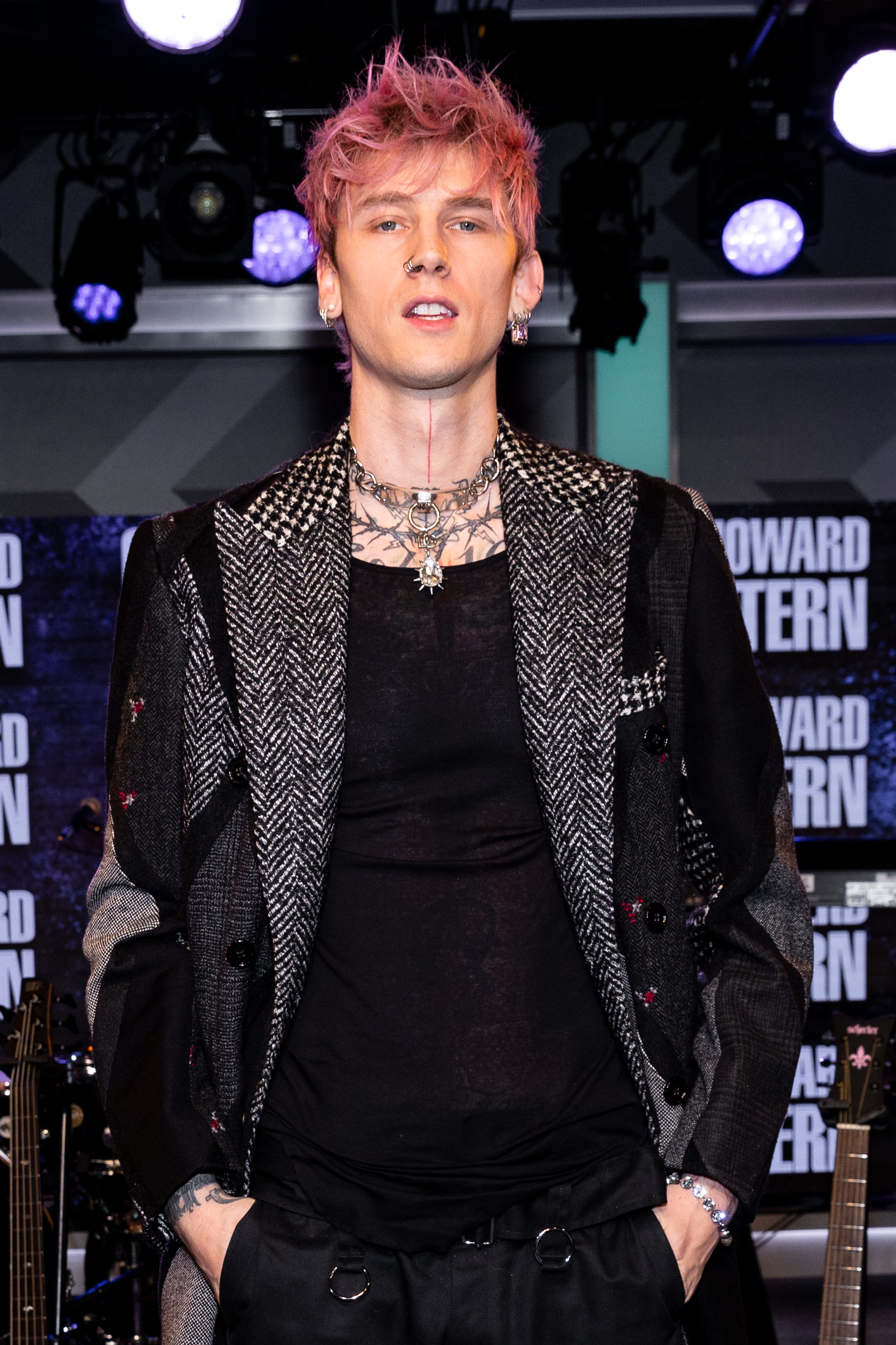 MGK with pink hair, wearing a studded jacket and necklace, standing before a microphone stand