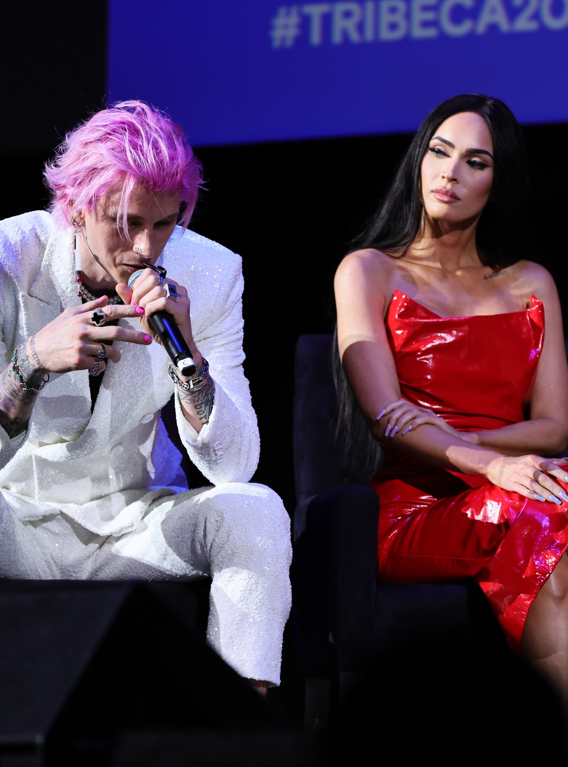 Musician in a white outfit and a woman in a shiny red dress sitting on stage, both with microphones. #TRIBECA2022 sign in view
