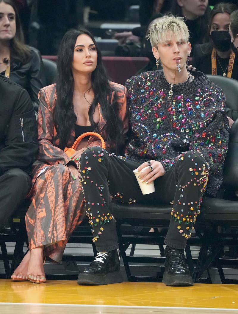 Meganin a striped outfit and Machine Gun Kelly in a sparkly top sit side by side at a sports event