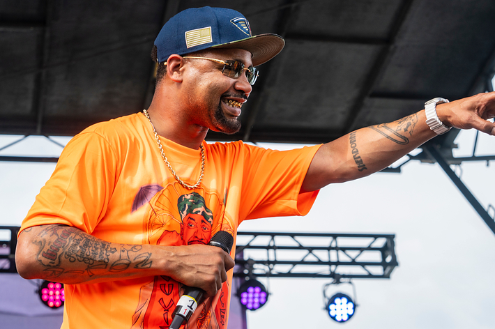 Juvenile on stage performing with microphone, wearing an orange shirt and a blue cap