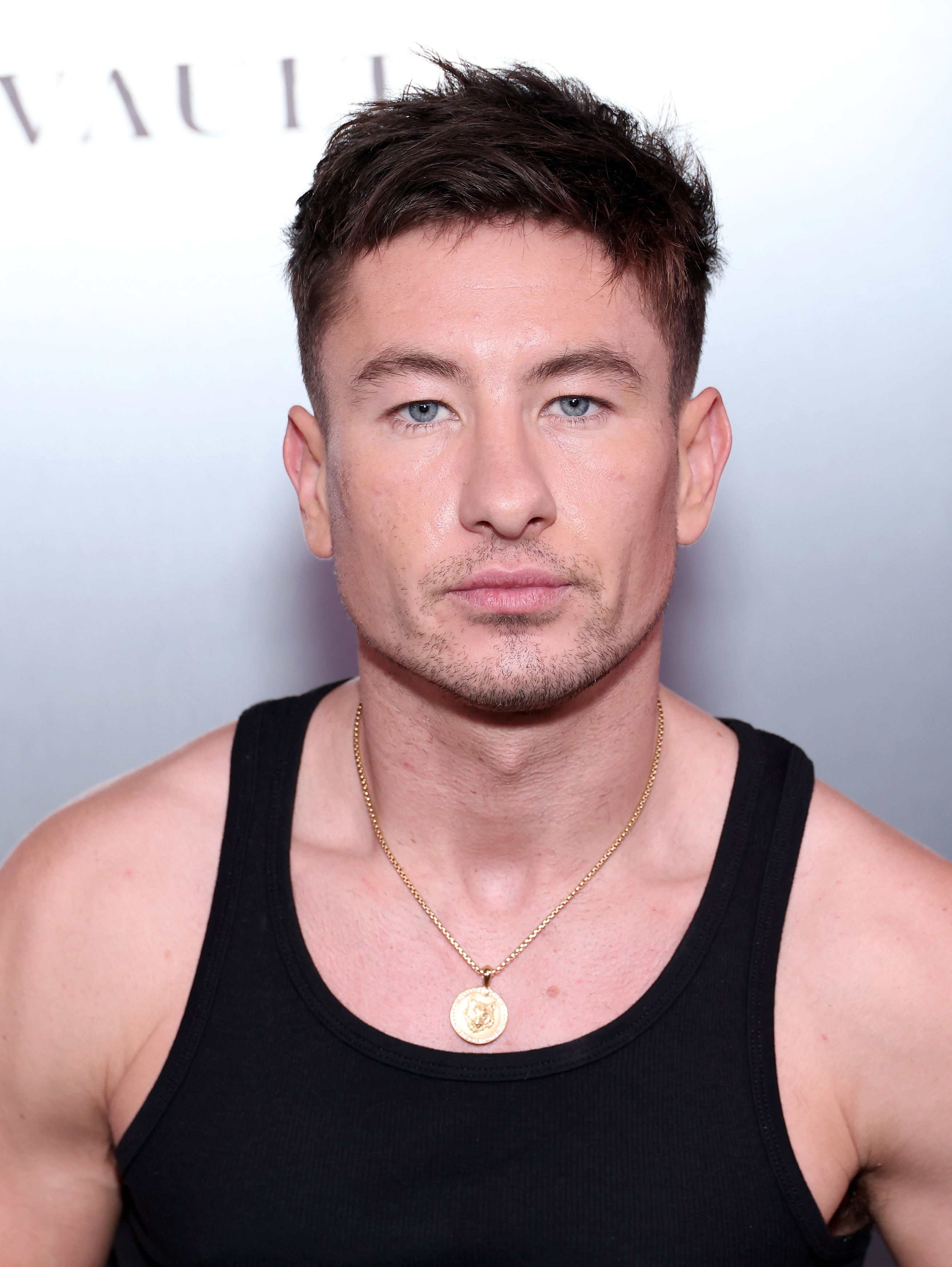 Barry Keoghan in a black sleeveless top with a gold necklace, posing in front of a promotional backdrop