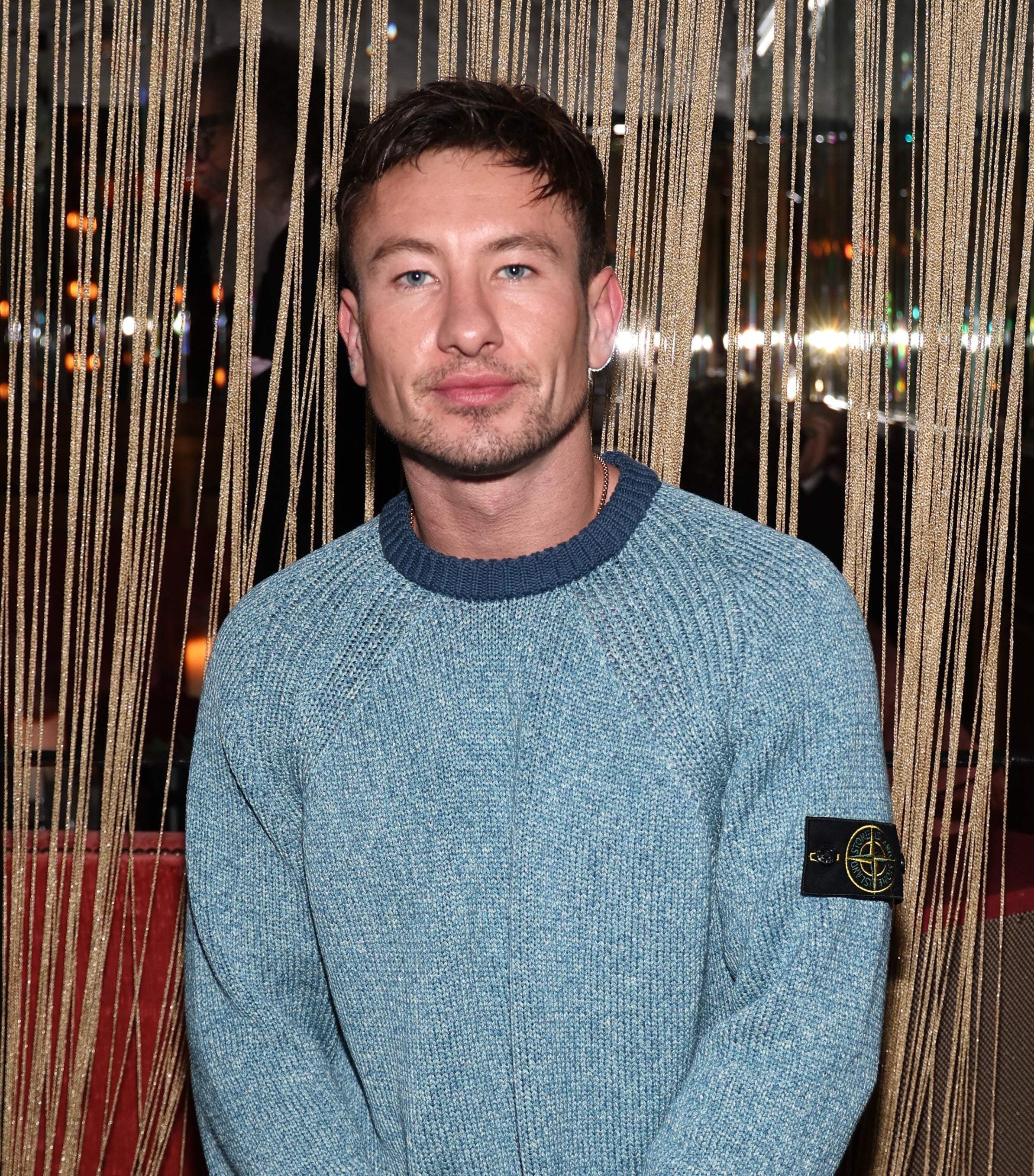 Barry Keoghan in a teal sweater and navy trousers posing with hands clasped