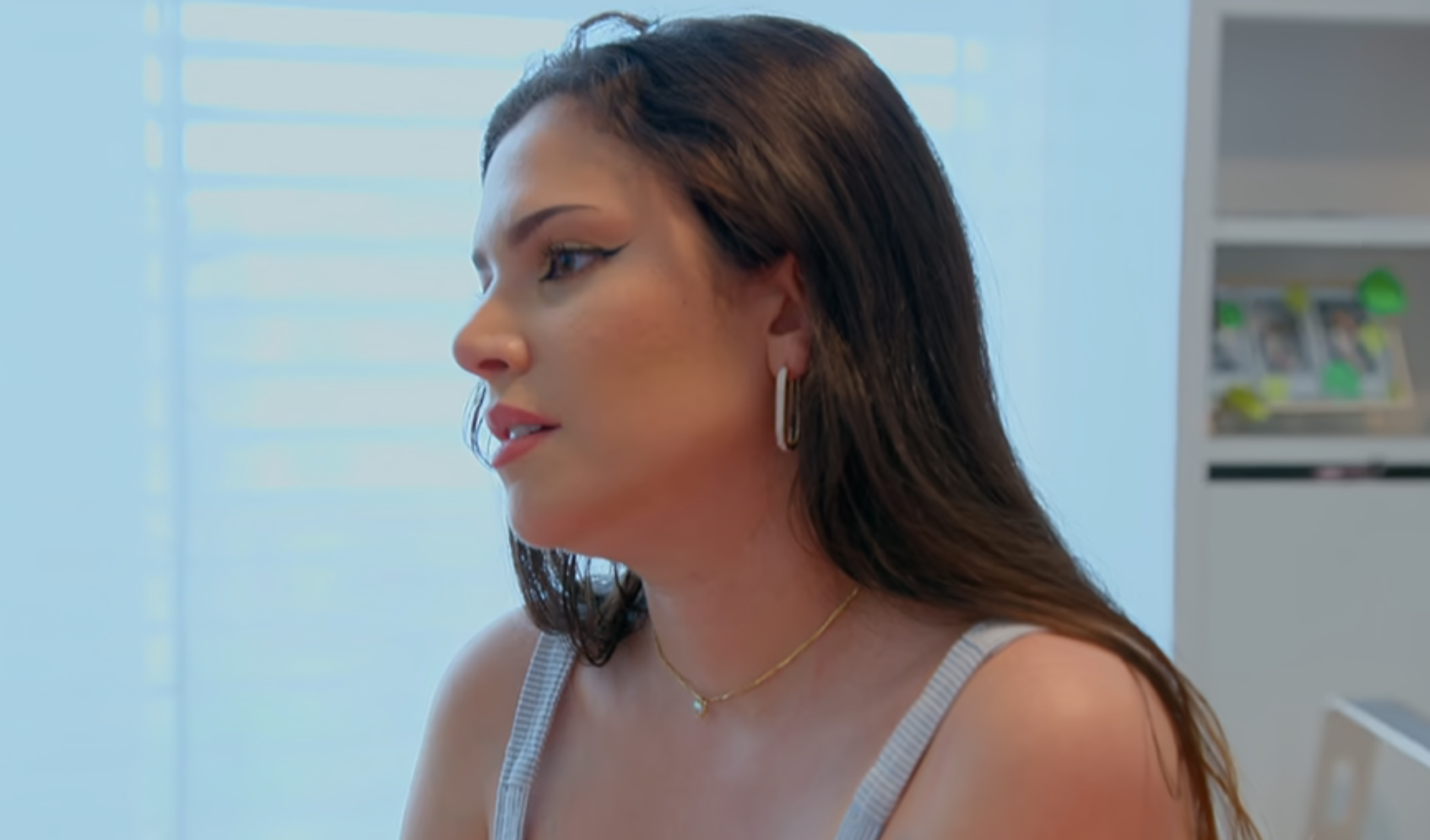 Woman in a sleeveless top with hoop earrings, sitting indoors, looking to the side, with a thoughtful expression