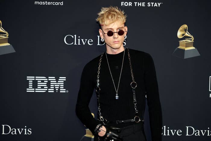 Machine Gun Kelly posing in a black sweater and sunglasses at an event with sponsor logos and Grammy statues in the background