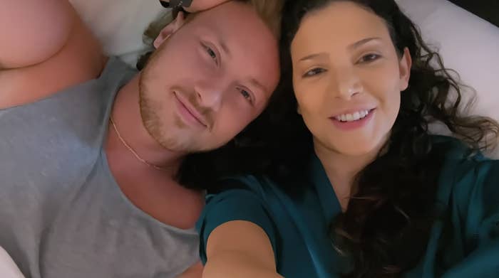 Two people smiling while lying down, close-up selfie view. No identifiable persons named