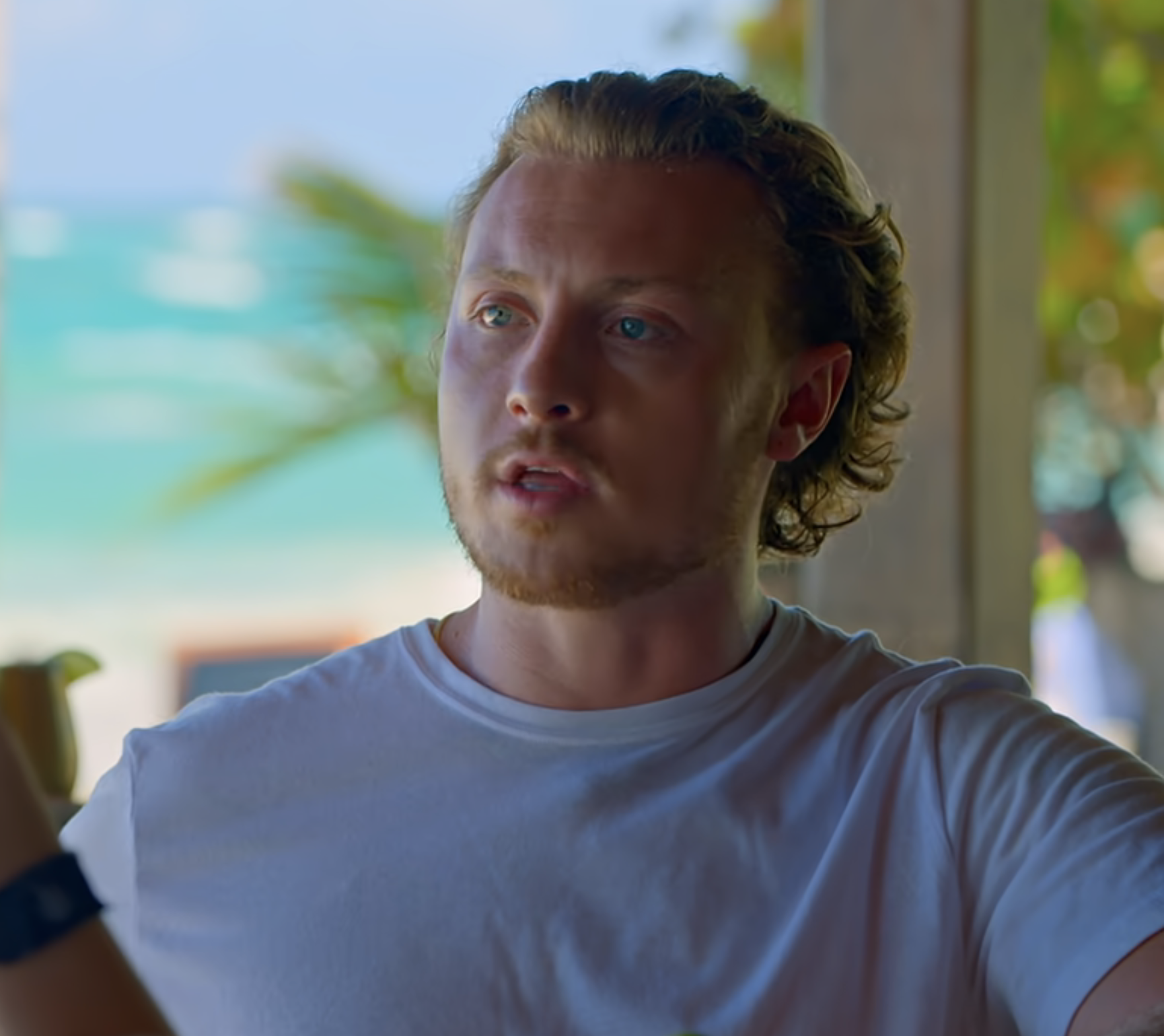 Johnny in a T-shirt and talking to another whose reflection is seen in a mirror, with a tropical backdrop visible through a window