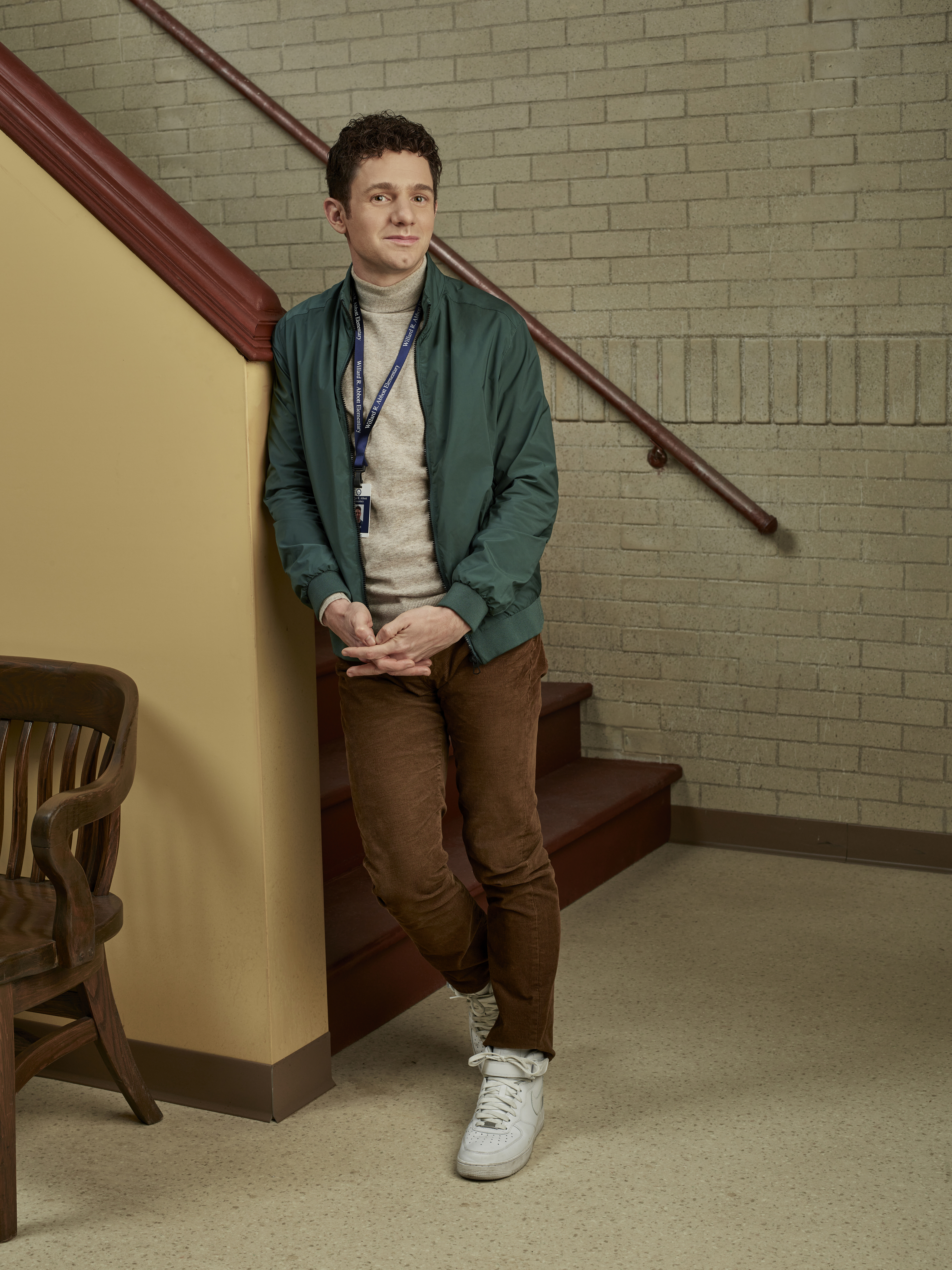Man leaning against wall by stairs, wearing a green jacket, brown pants, and white sneakers