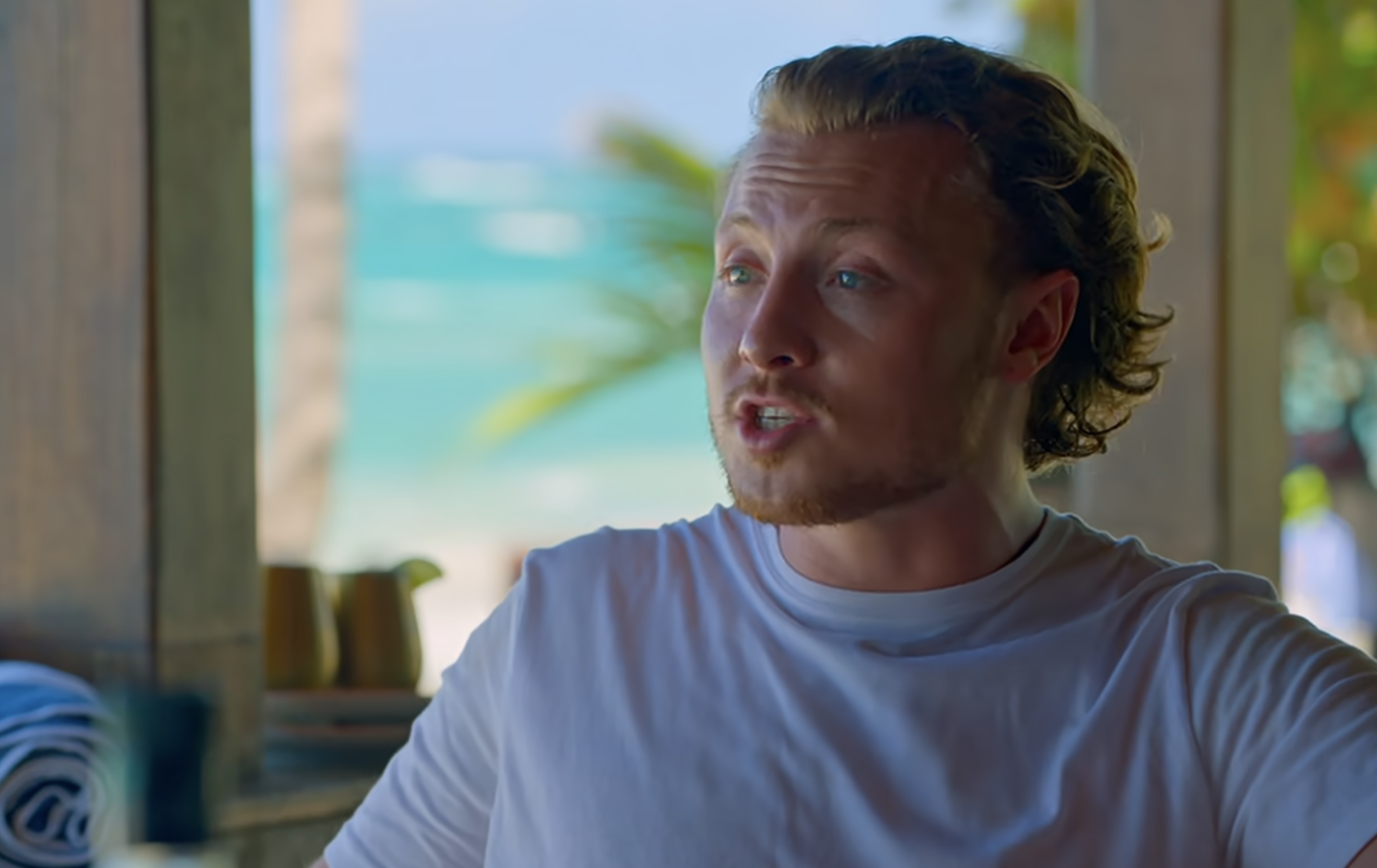 Man in casual white tee sitting indoors with ocean view outside window