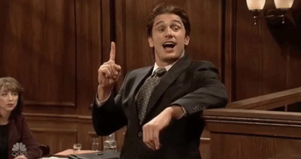 Man in courtroom scene raising his finger, in a suit, appears to be making a point