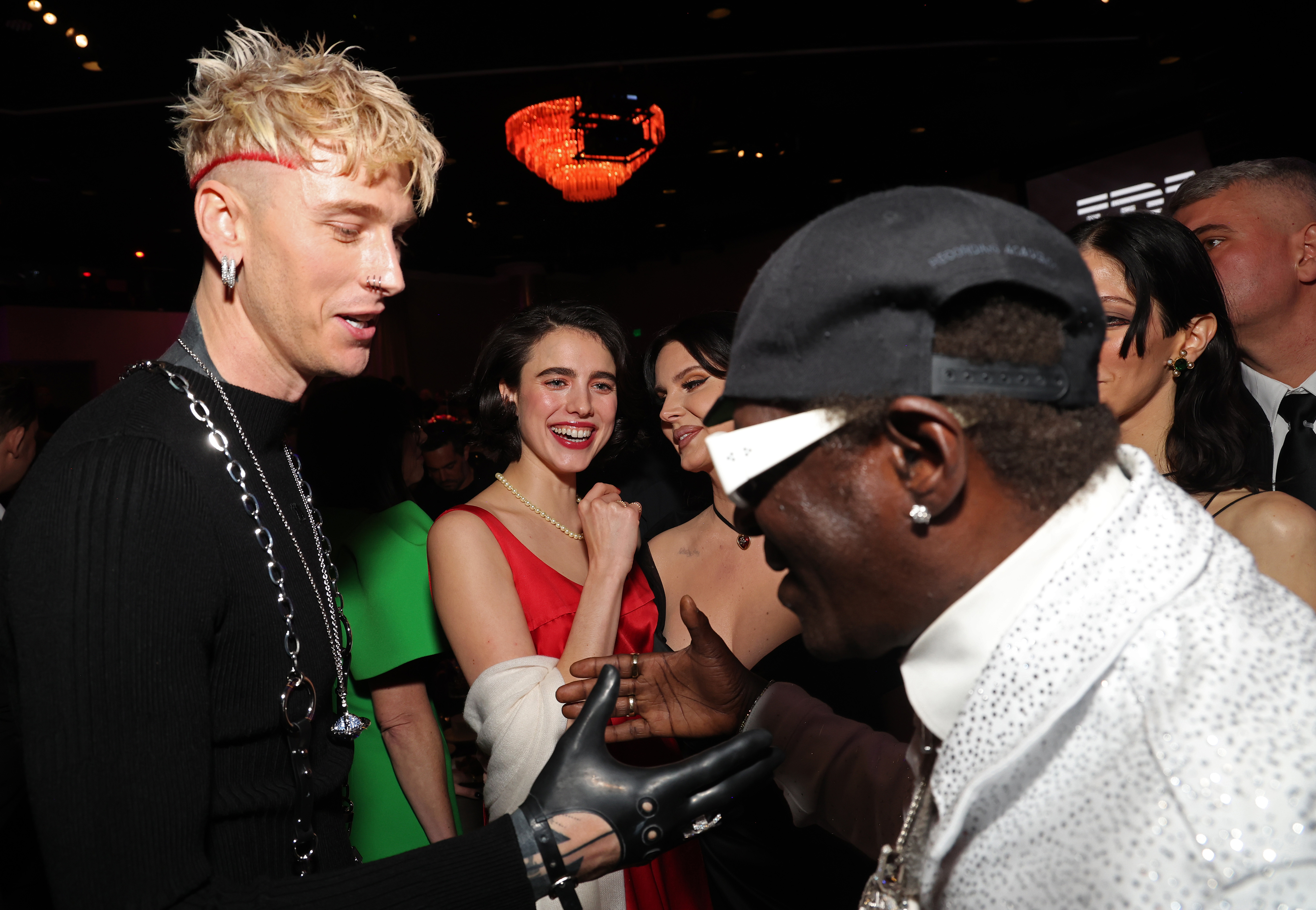 Machine Gun Kelly in a punk-inspired outfit talking with Bobby Brown, who&#x27;s wearing a sparkling jacket, at an event. Others present