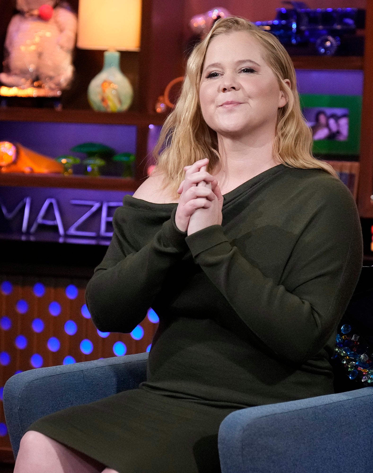Amy Schumer in a TV show setting wears an off-shoulder dress, seated, smiling