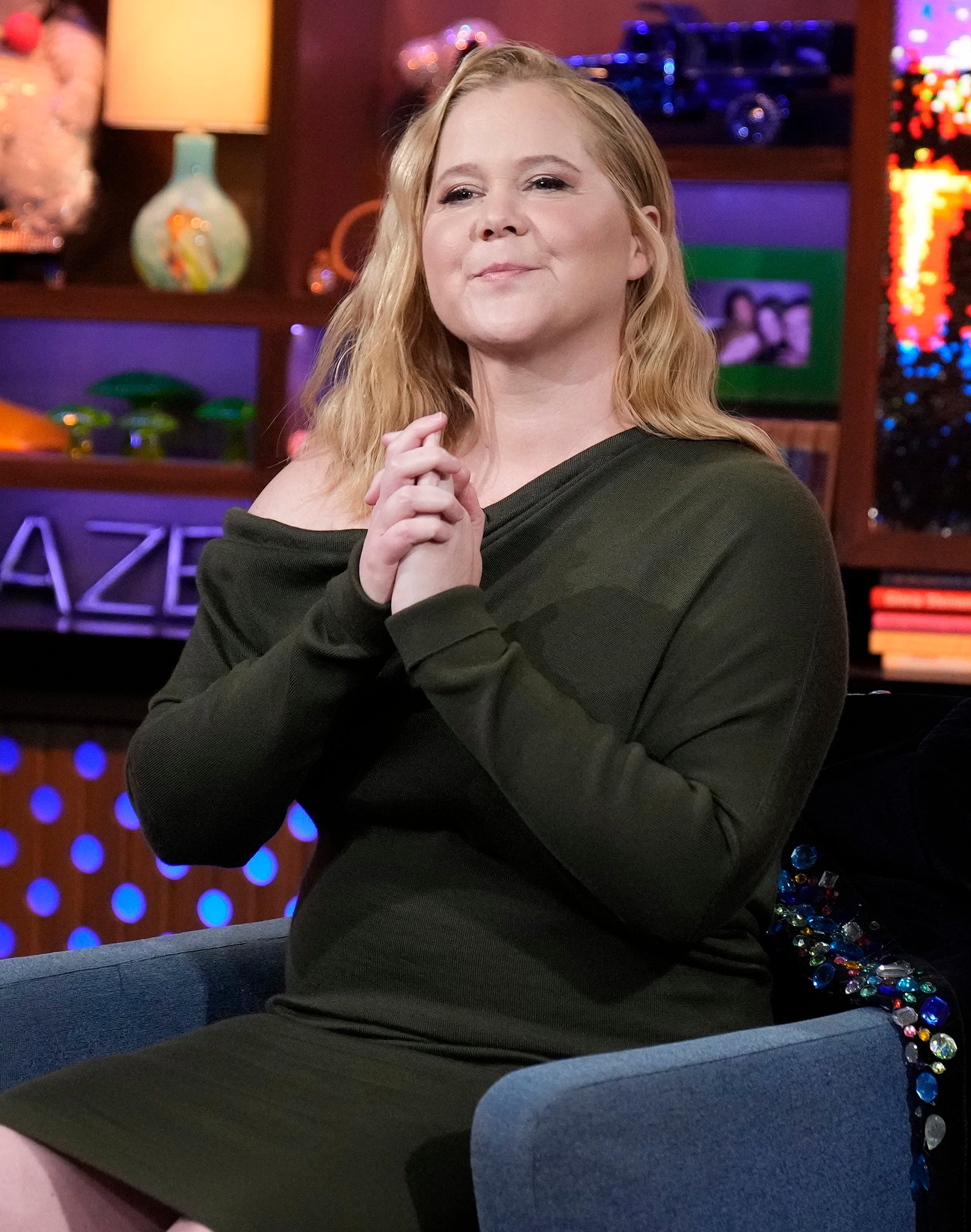 Amy Schumer in a TV show setting wears an off-shoulder dress, seated, smiling