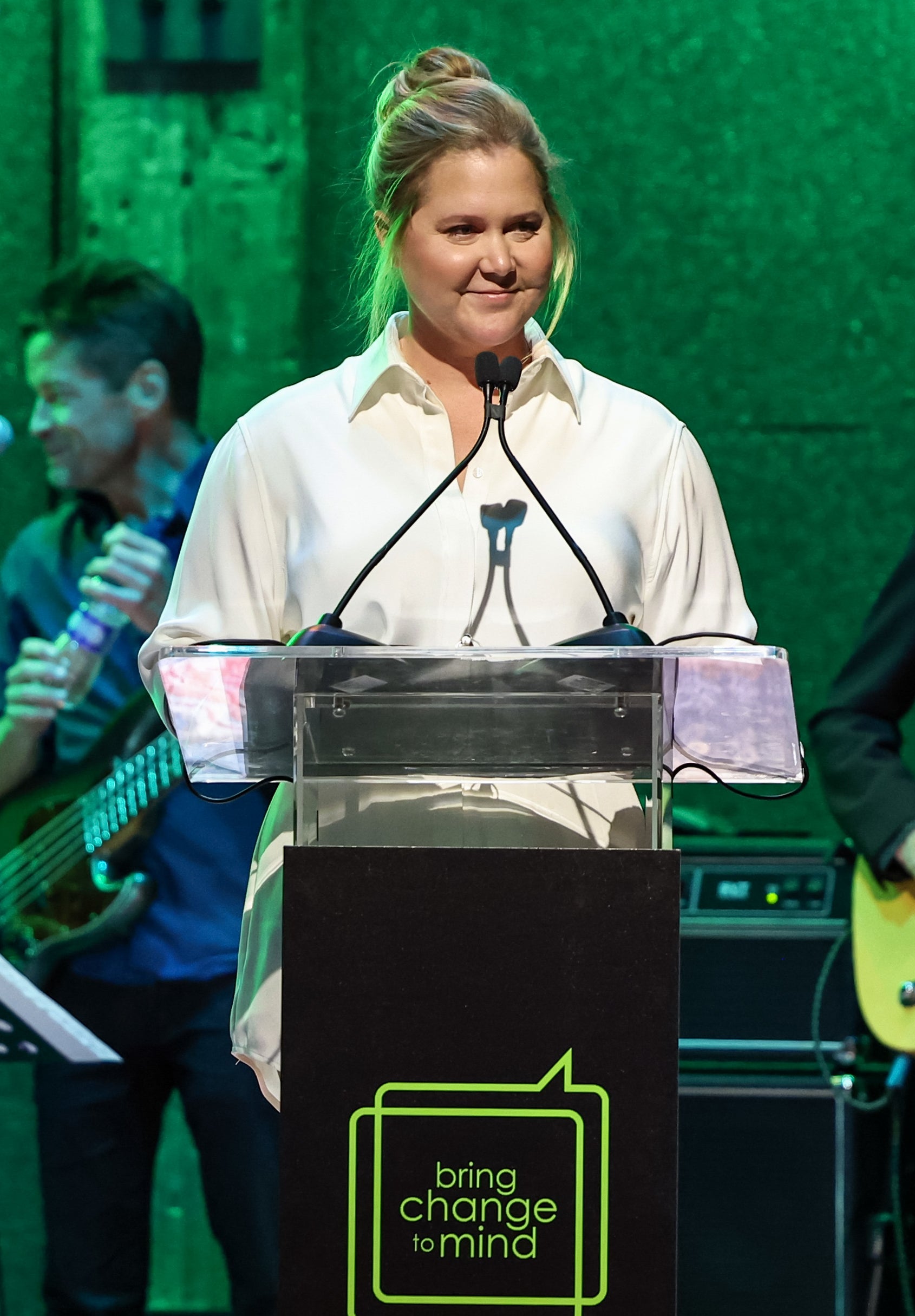 Amy Schumer stands at a podium with "bring change to mind" sign, musicians in background. She wears a white blouse