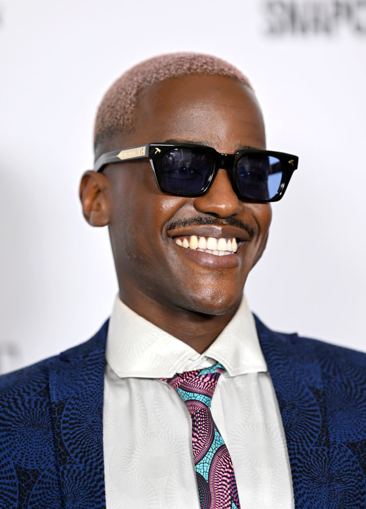 Ncuti in patterned suit and tie with sunglasses smiling at an event