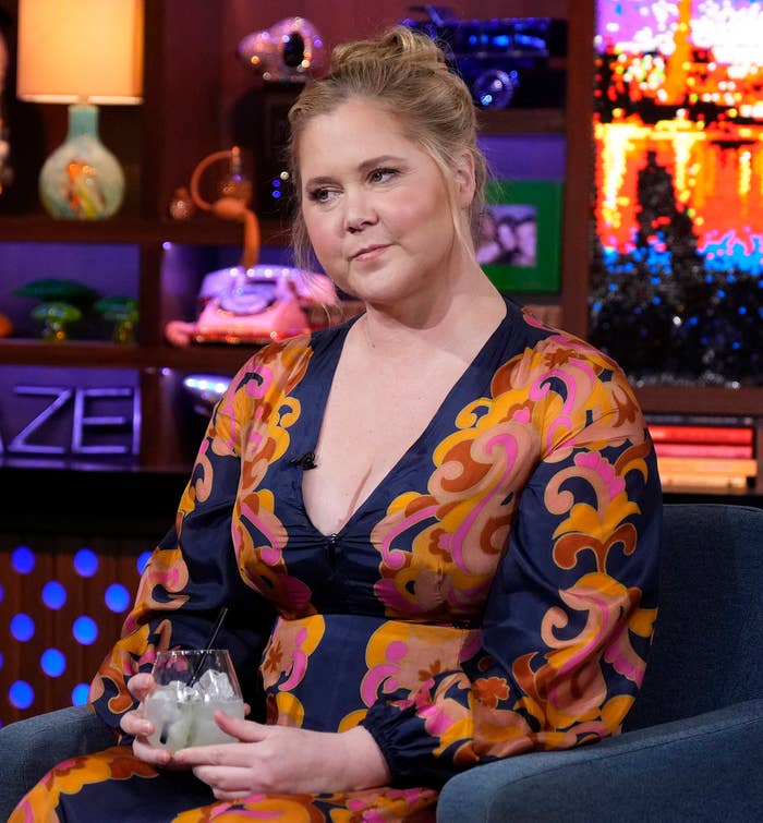 Amy Schumer wearing a patterned dress, seated, holding a glass, on a talk show set
