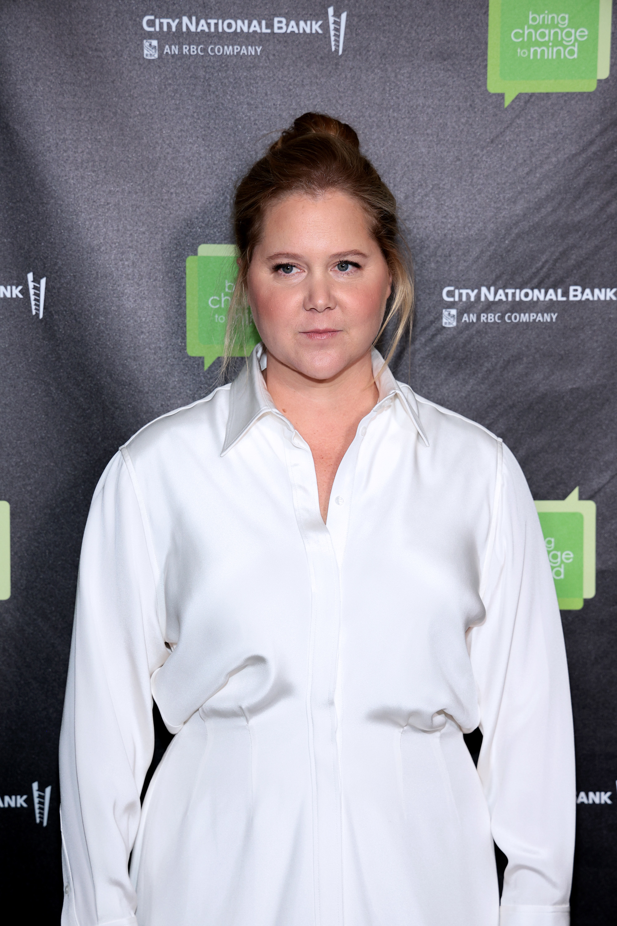 Amy Schumer wearing a white button-up shirt at an event with sponsor banners in the background