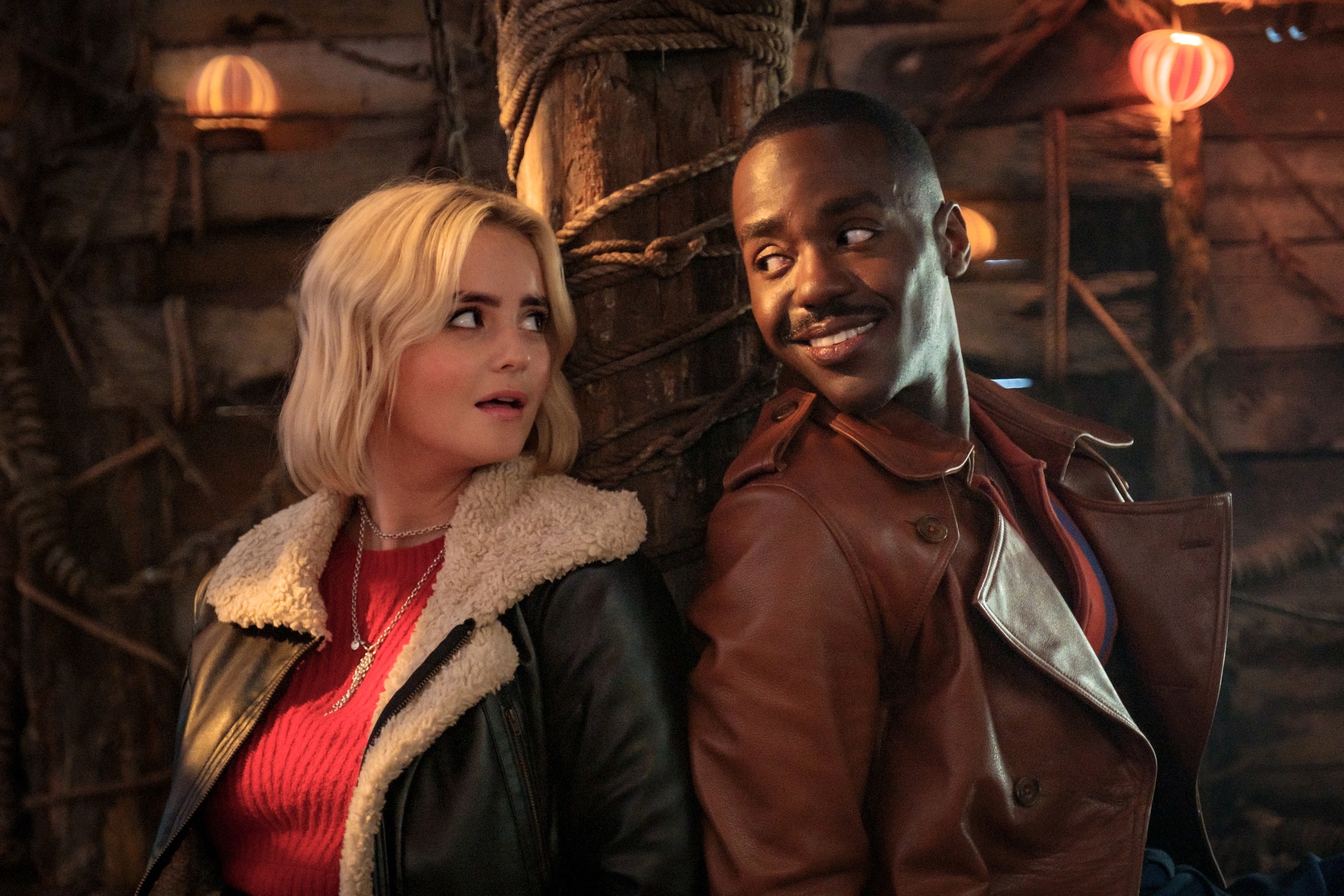 Ruby in a shearling jacket over top and the Doctor in a leather jacket, both smiling, in a warmly lit setting