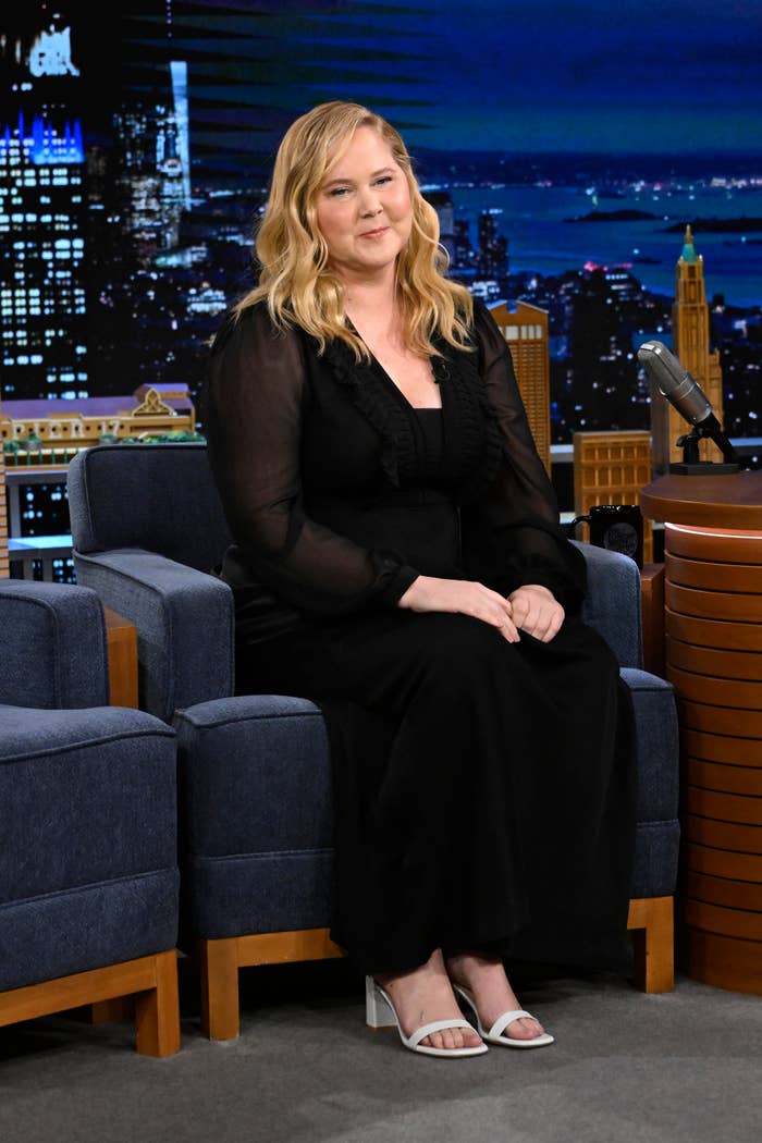 Amy Schumer dressed in a black outfit with sheer sleeves, seated on a talk show set