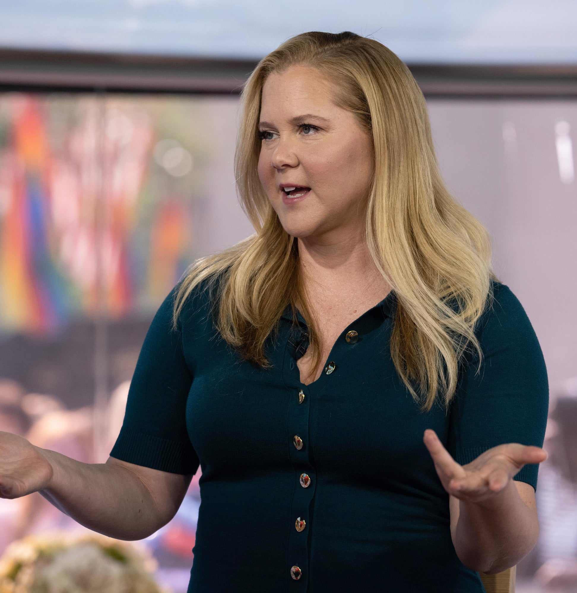 Amy Schumer in a casual outfit gestures while speaking, with blurry background