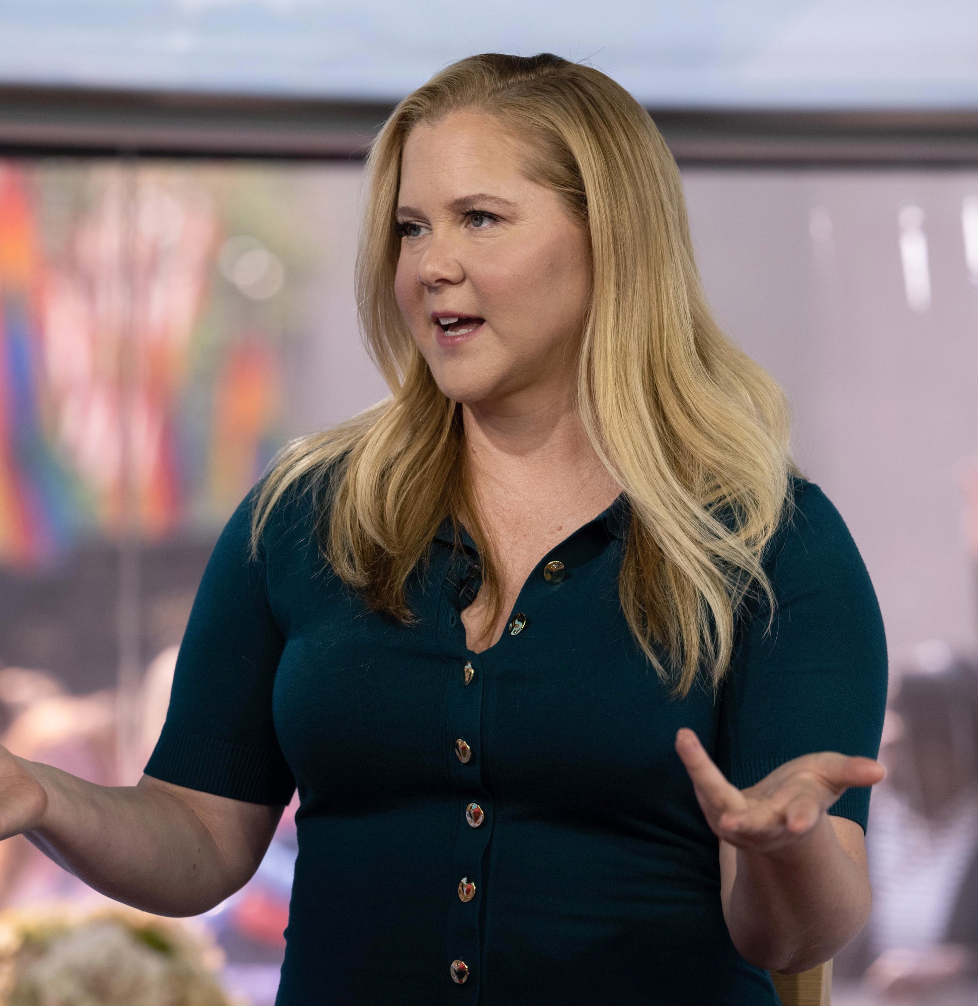 Amy Schumer in a casual outfit gestures while speaking, with blurry background