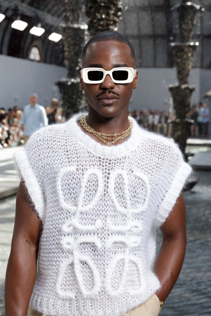 Ncuti wearing textured top with layered gold necklaces and sunglasses, standing in front of an audience