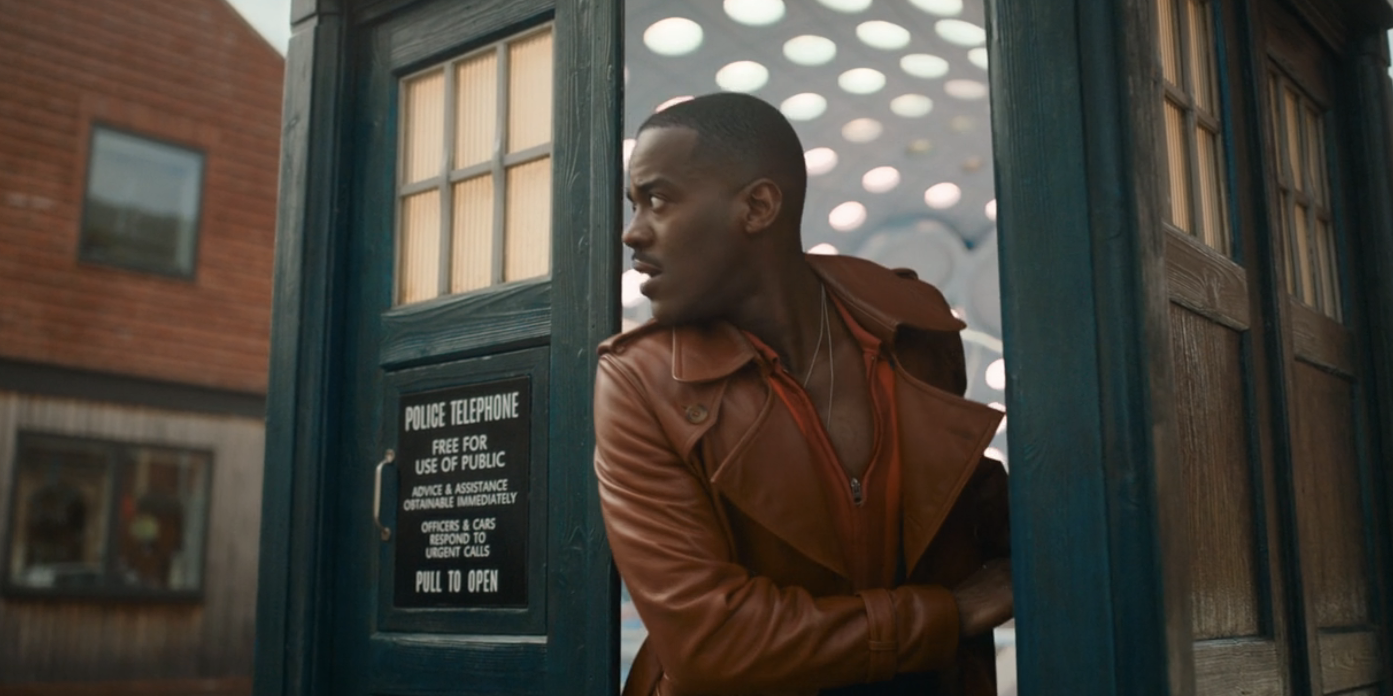 the Doctor in a leather jacket beside a British police box on a city street