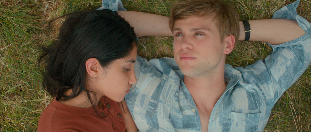 Two actors lie on grass in a scene, man in plaid, woman in a solid top, both gazing ahead