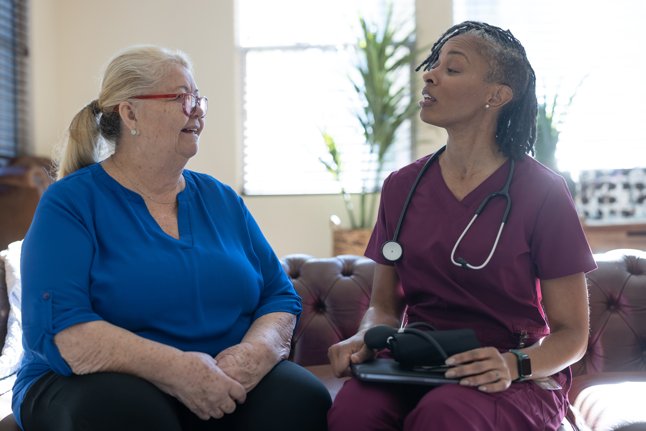 Healthcare professional in scrubs with stethoscope speaking to an older woman sitting indoors