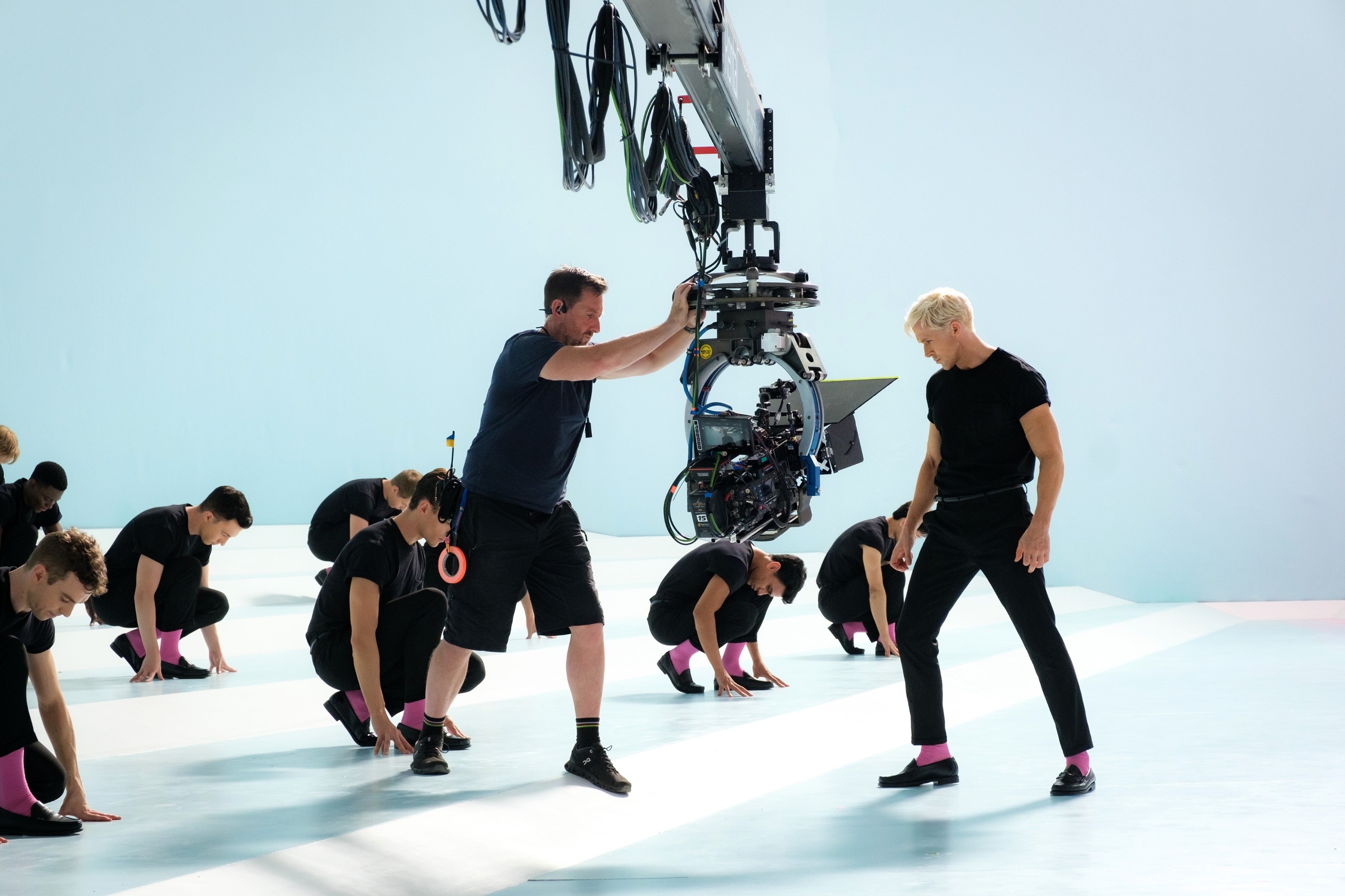 Crew filming a dance scene with a cameraman operating equipment and dancers in synchronised poses in the background
