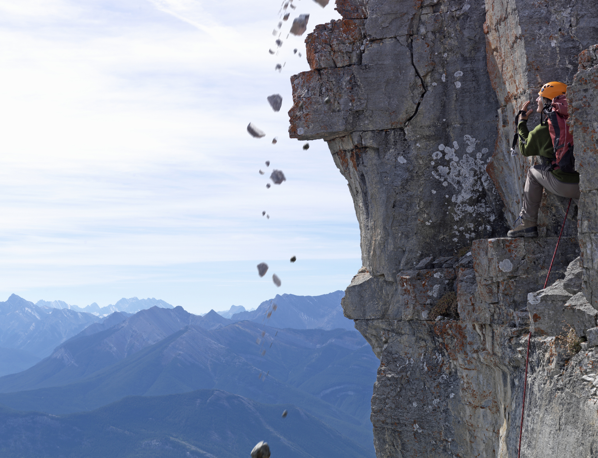 Climber secured with ropes on a mountain face, rocks falling beside them, expansive view of peaks in the background