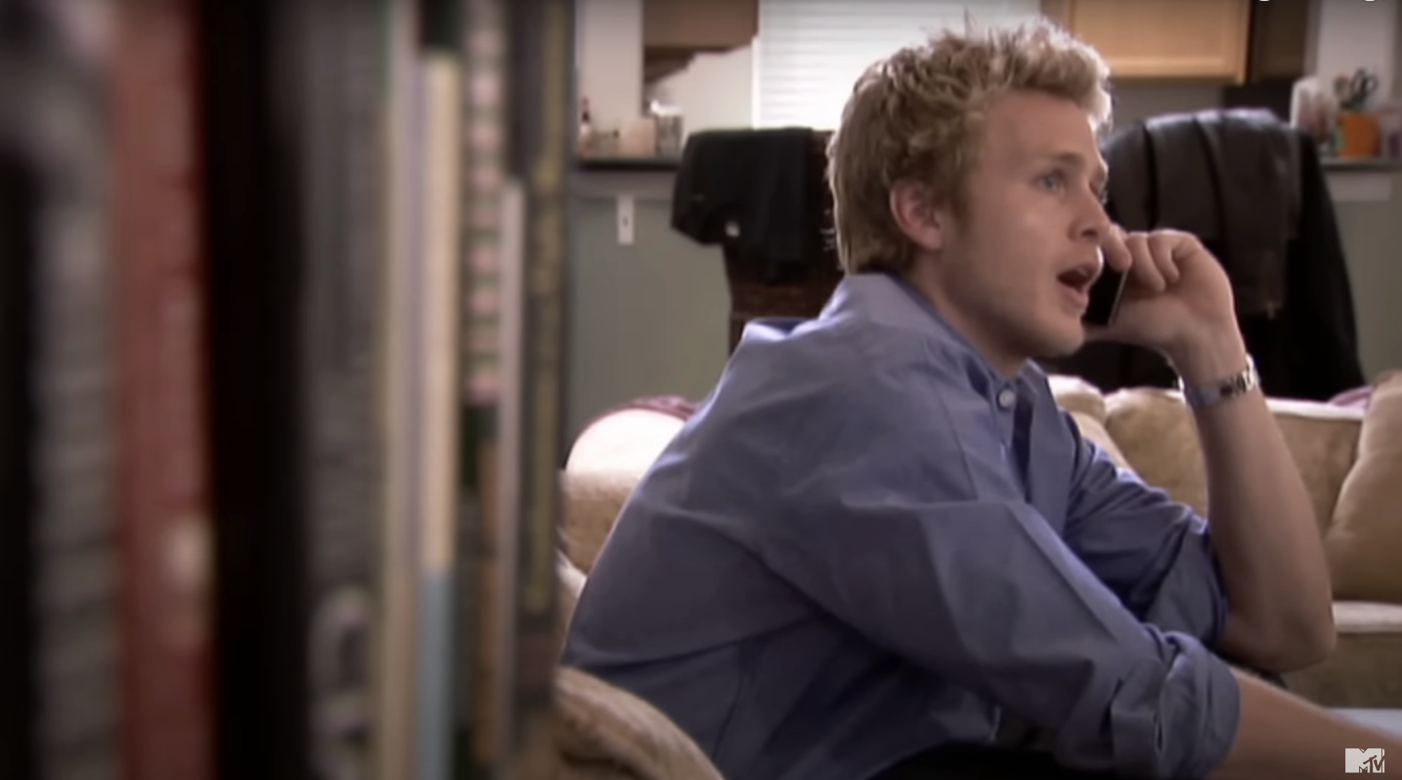 Spencer sitting on a couch talking on the phone, appearing surprised, in a casual setting