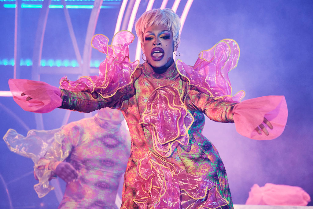 Yvie in a shimmery outfit with voluminous sleeves on stage, expressing with hands outstretched