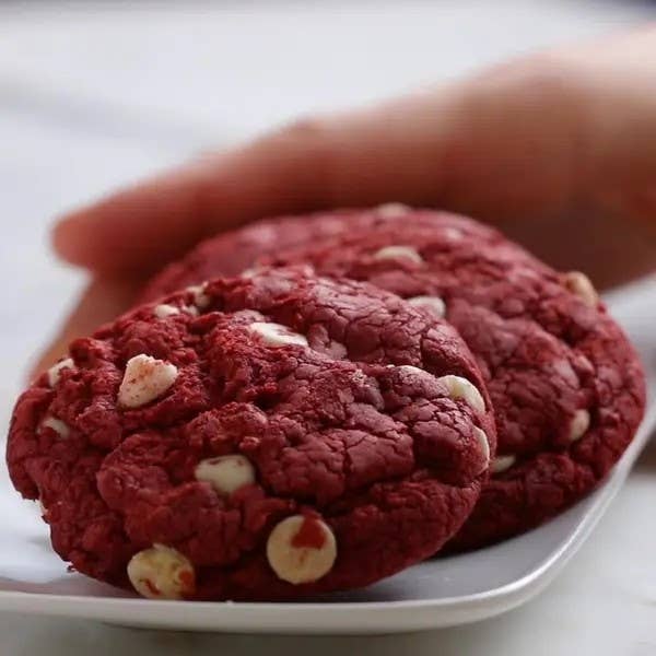 A hand reaching for a red velvet cookie with white chocolate chips on a plate