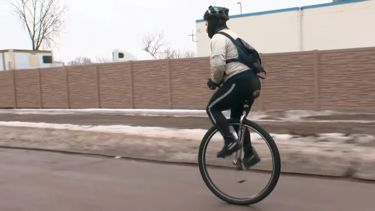 Person riding a unicycle on a roadside with snow patches and a blue wall in the background