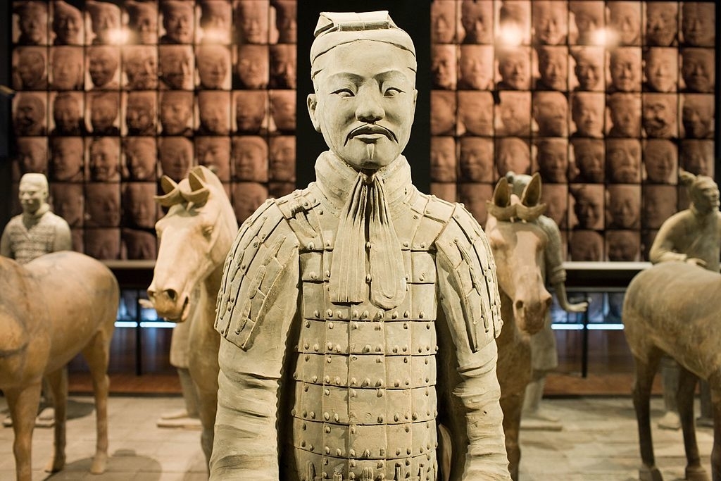 Replica of a Terracotta Warrior statue in front of a wall displaying many warrior faces