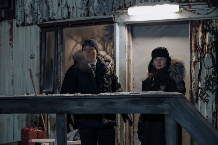 Two characters in winter gear stand by a cabin in a scene from a TV series
