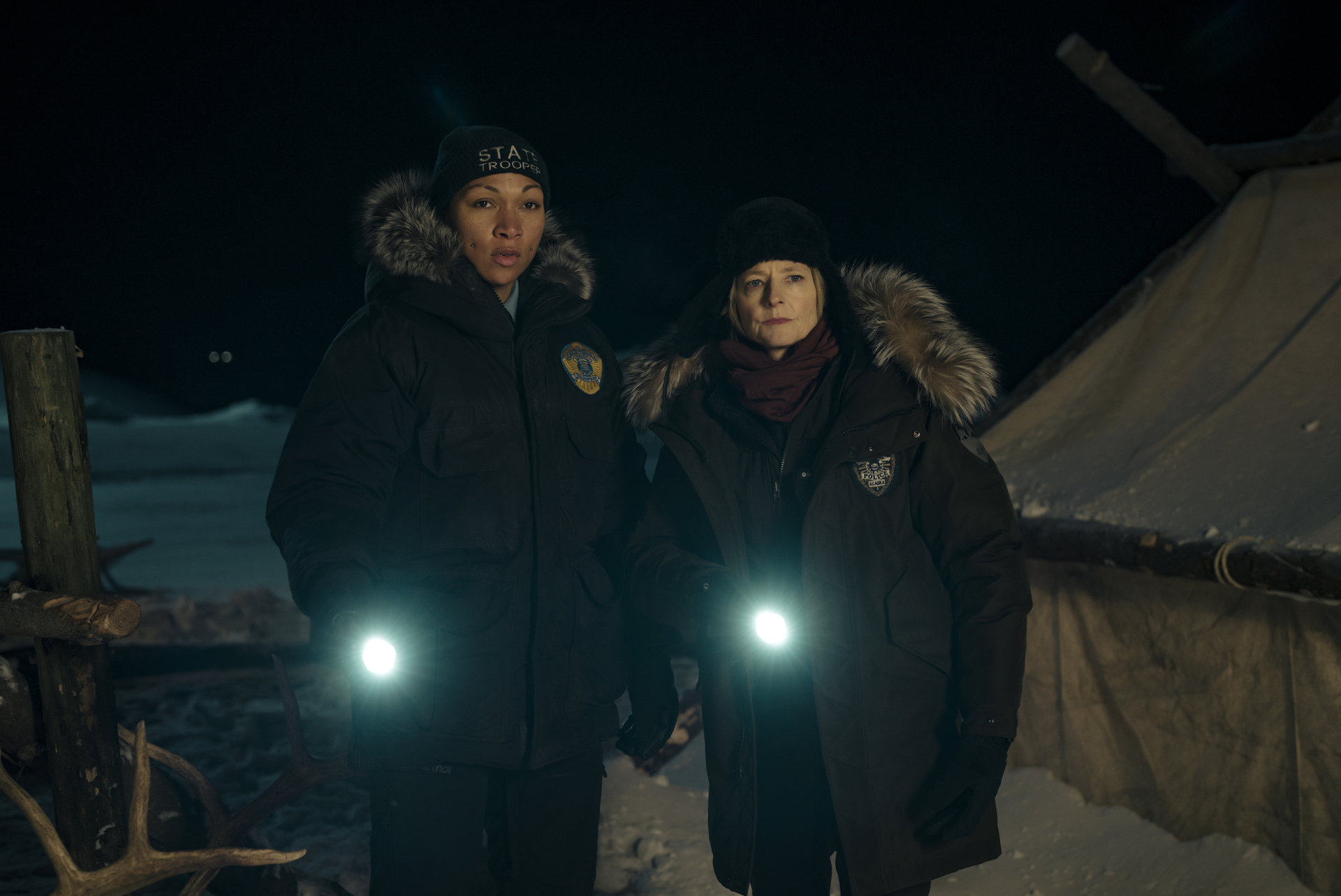 Two characters from a TV show wearing winter police uniforms, standing outside at night with flashlights