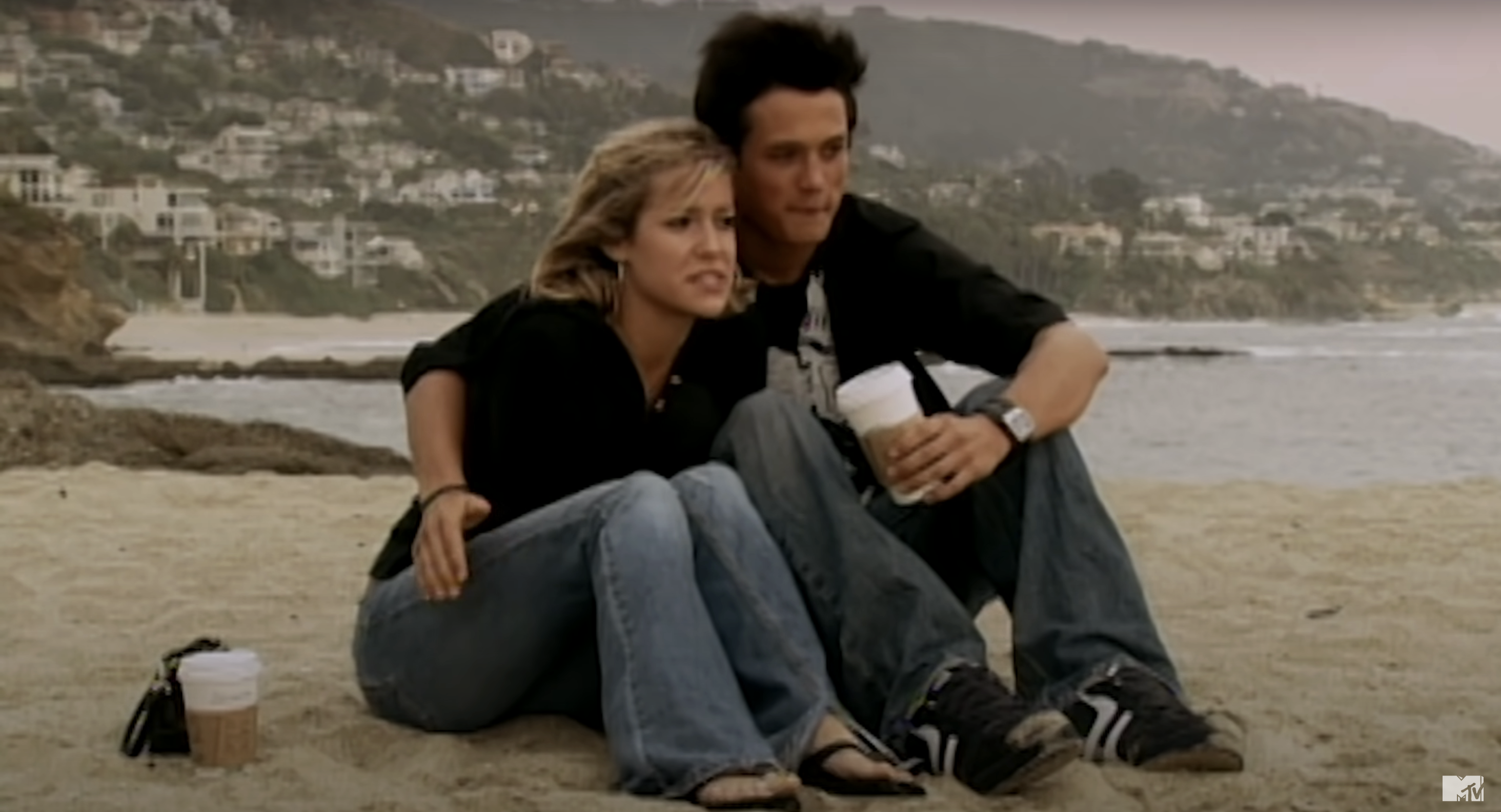 Stephen and Kristin sit closely together on a beach, gazing into the distance, dressed in casual attire