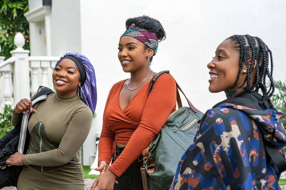 Three women smiling and walking together, one wearing a headscarf, one with a headband, and one with braided hair
