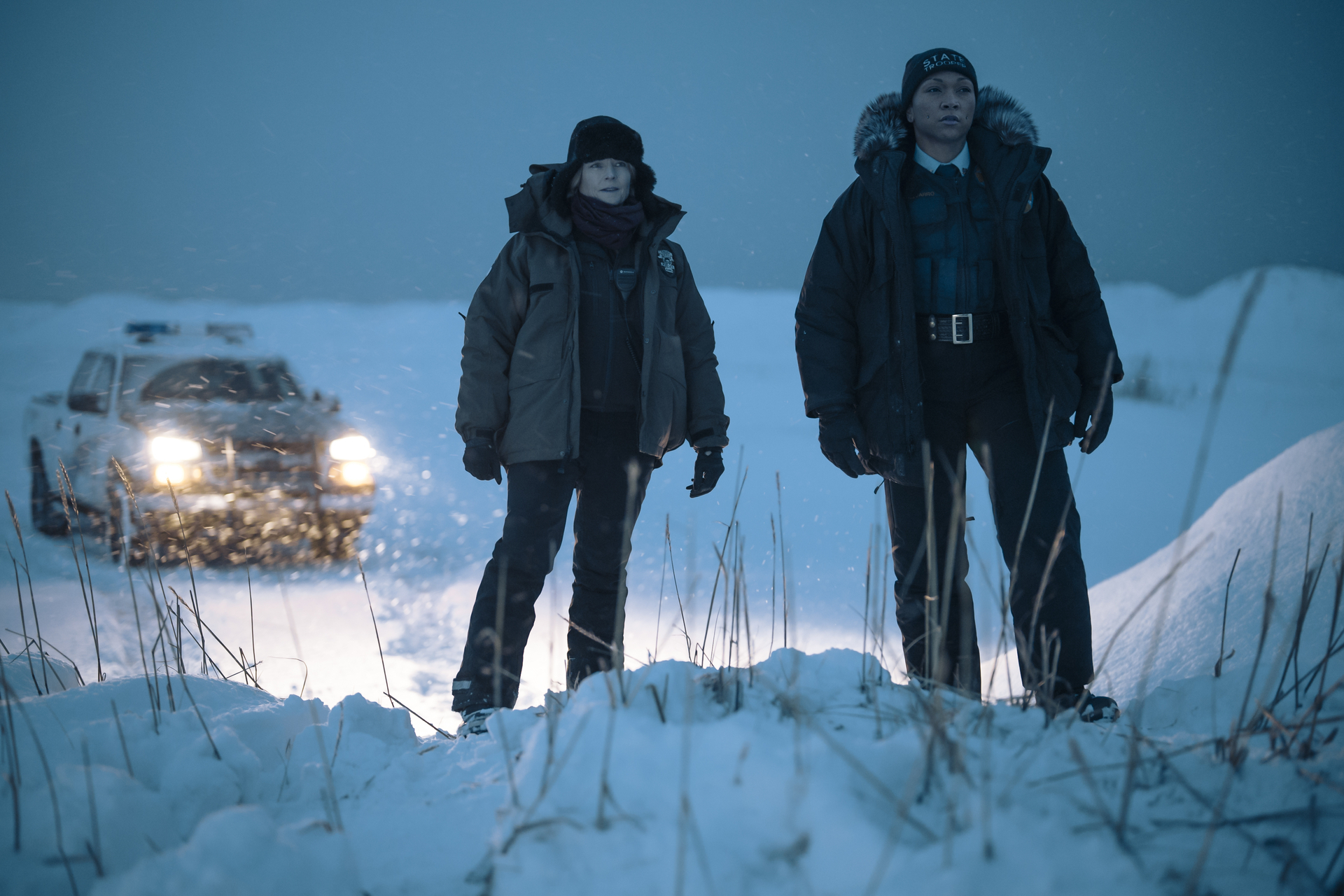 Two characters in heavy winter gear stand in the snow with a vehicle behind them, in a scene from a TV show or movie