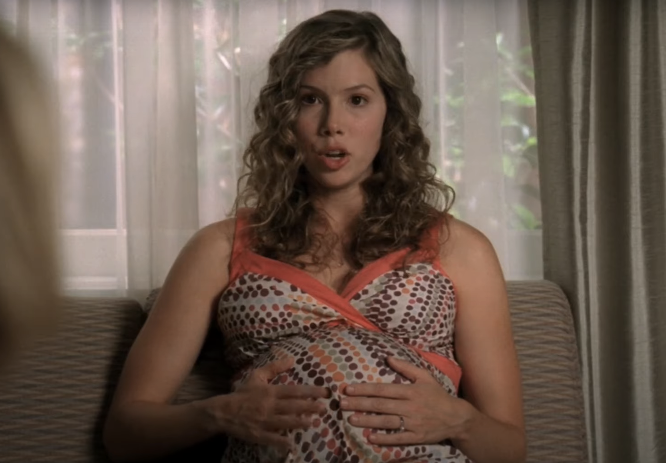 A woman in a patterned top gesturing with hands on her belly, sitting indoors
