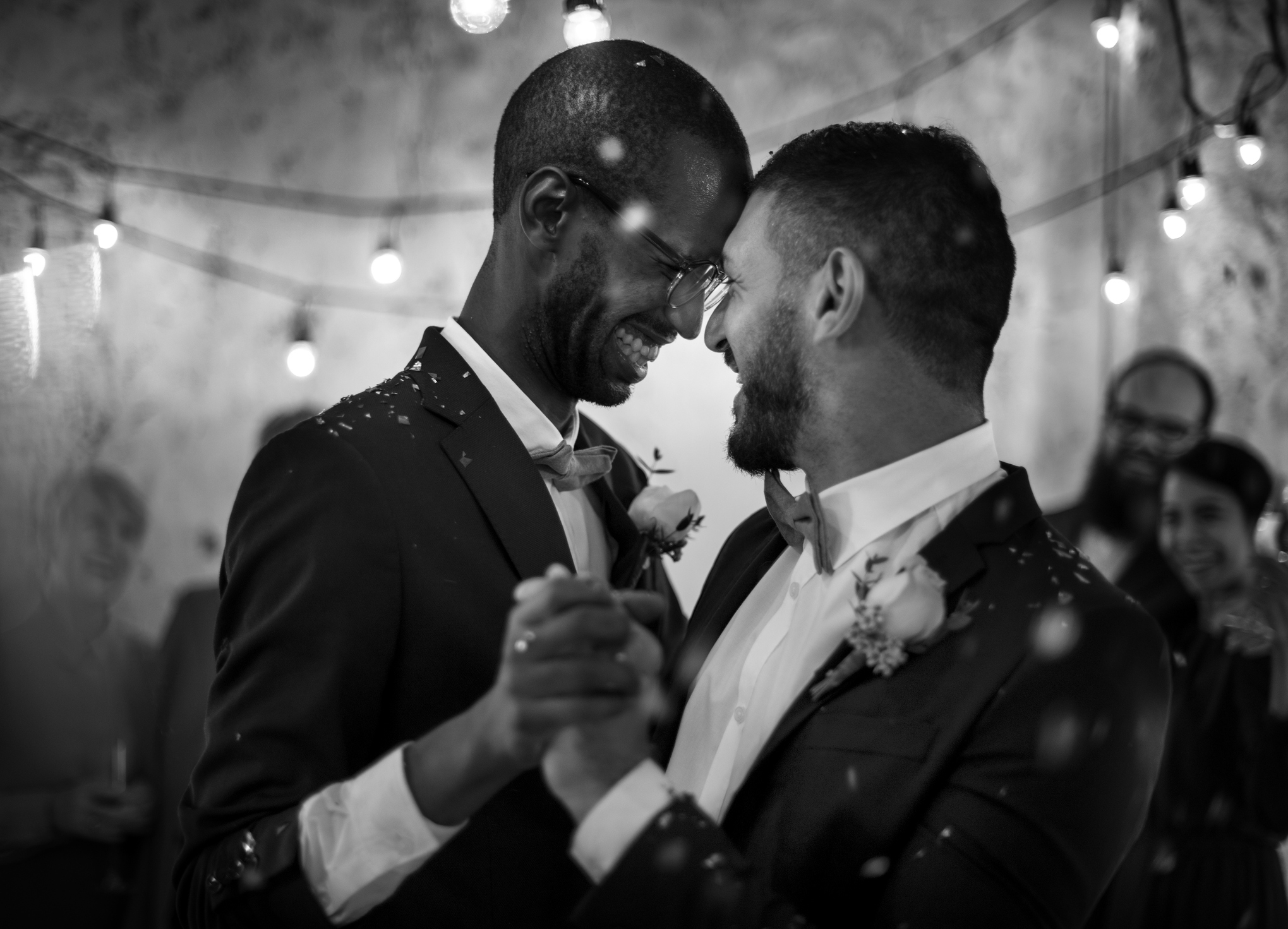Two grooms smiling at each other, sharing a dance, guests in background
