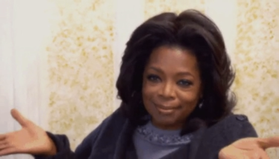 Oprah Winfrey smiling, palms up, in a gray sweater, expressing a welcoming gesture