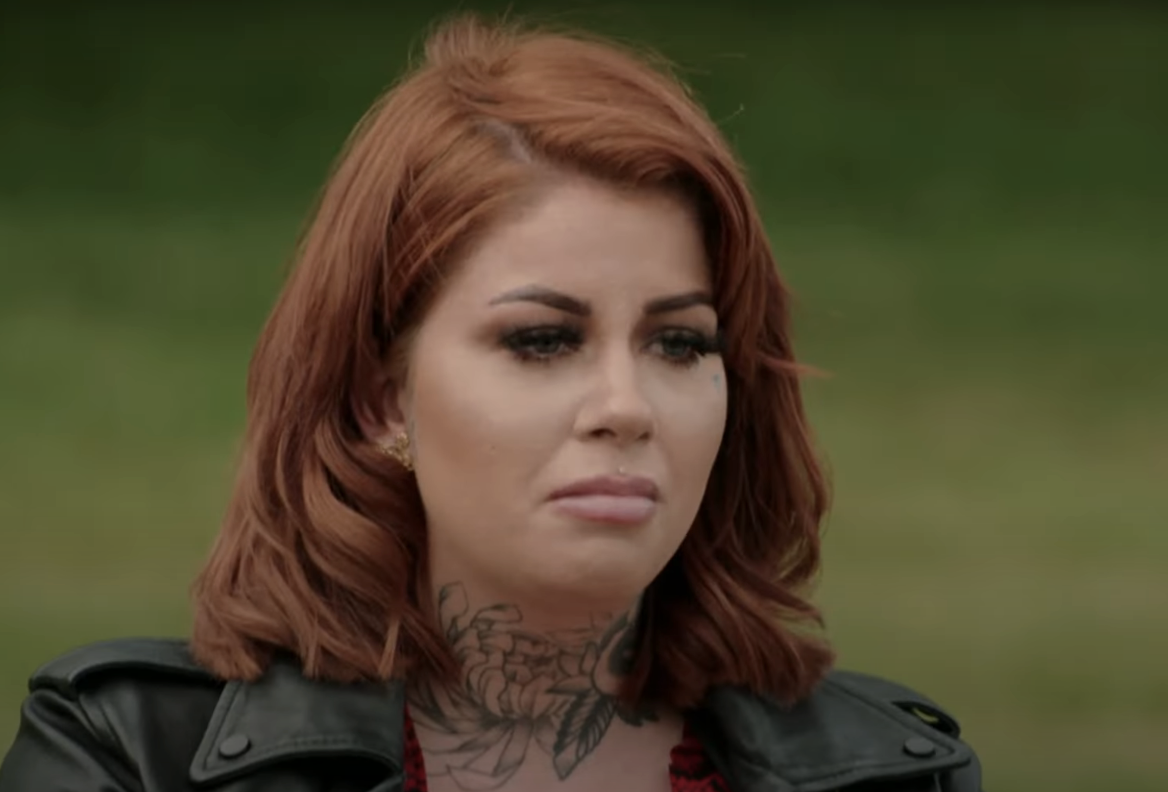 Close-up of Gemma looking pensive, wearing a dark jacket with a visible tattoo on her chest