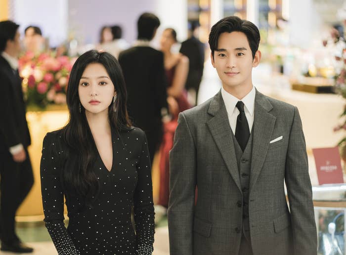 Dos personajes de K-drama en evento elegante, mujer con vestido escotado y hombre con traje de rayas